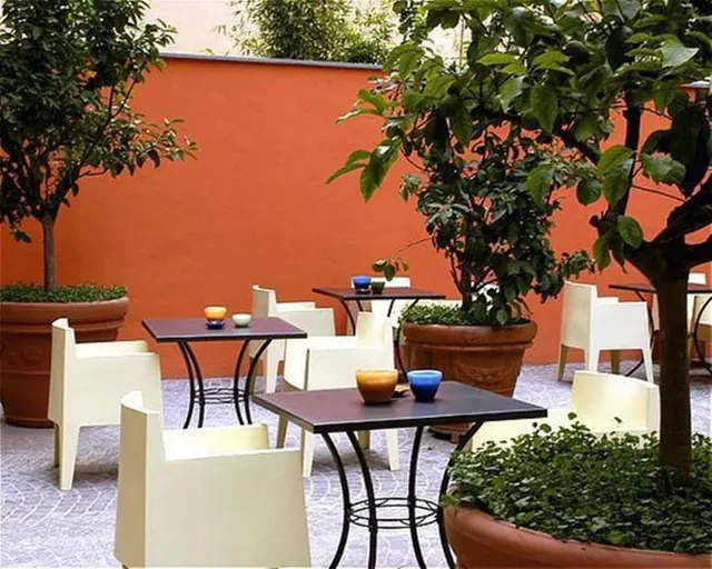 An outdoor courtyard terrace with white chairs, dark square tables, potted citrus trees and an orange wall.