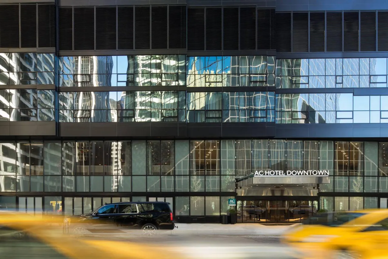 Glass facade of the AC Hotel Downtown with its entrance canopy, a black SUV parked at the kerb and blurred yellow taxis in the foreground.