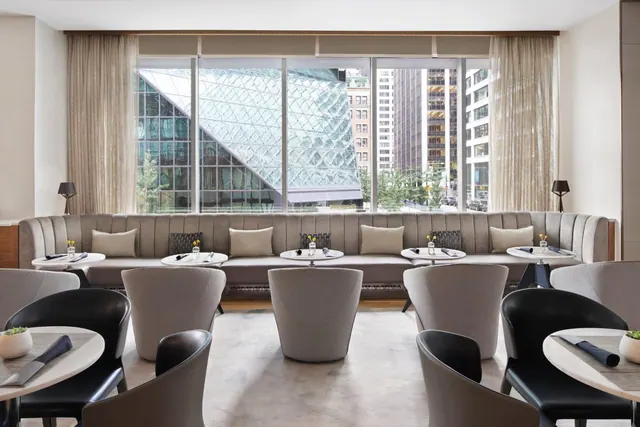 A restaurant with a long grey banquette, round tables and grey armchairs, with large windows showing a glass-faceted building and city towers outside.