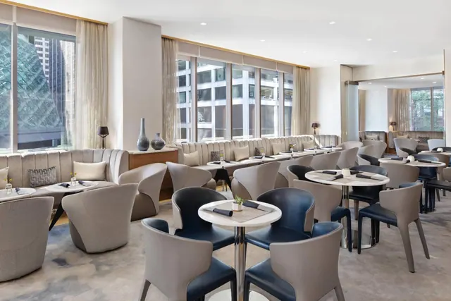 A restaurant dining room with upholstered banquette seating and round tables set with napkins, with tall windows looking onto nearby office buildings.