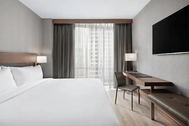 A bedroom with a made king bed, a wall-mounted television, a desk with a chair and a bench, and a window with sheer curtains overlooking tall buildings.