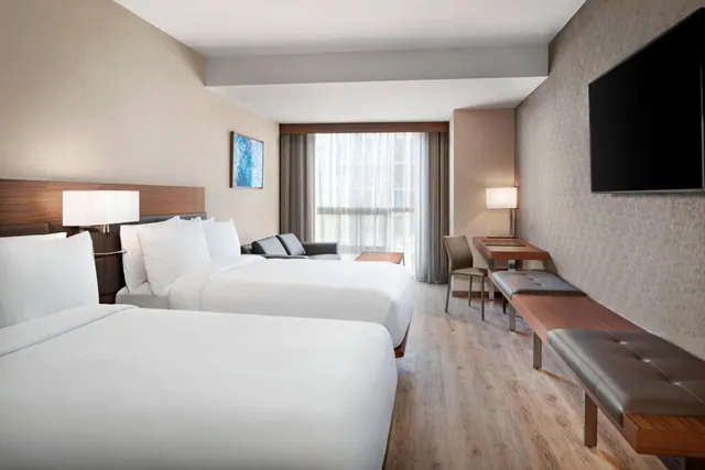 A hotel room with two double beds, a grey seating area, a wooden desk with a chair, and a wall-mounted television.