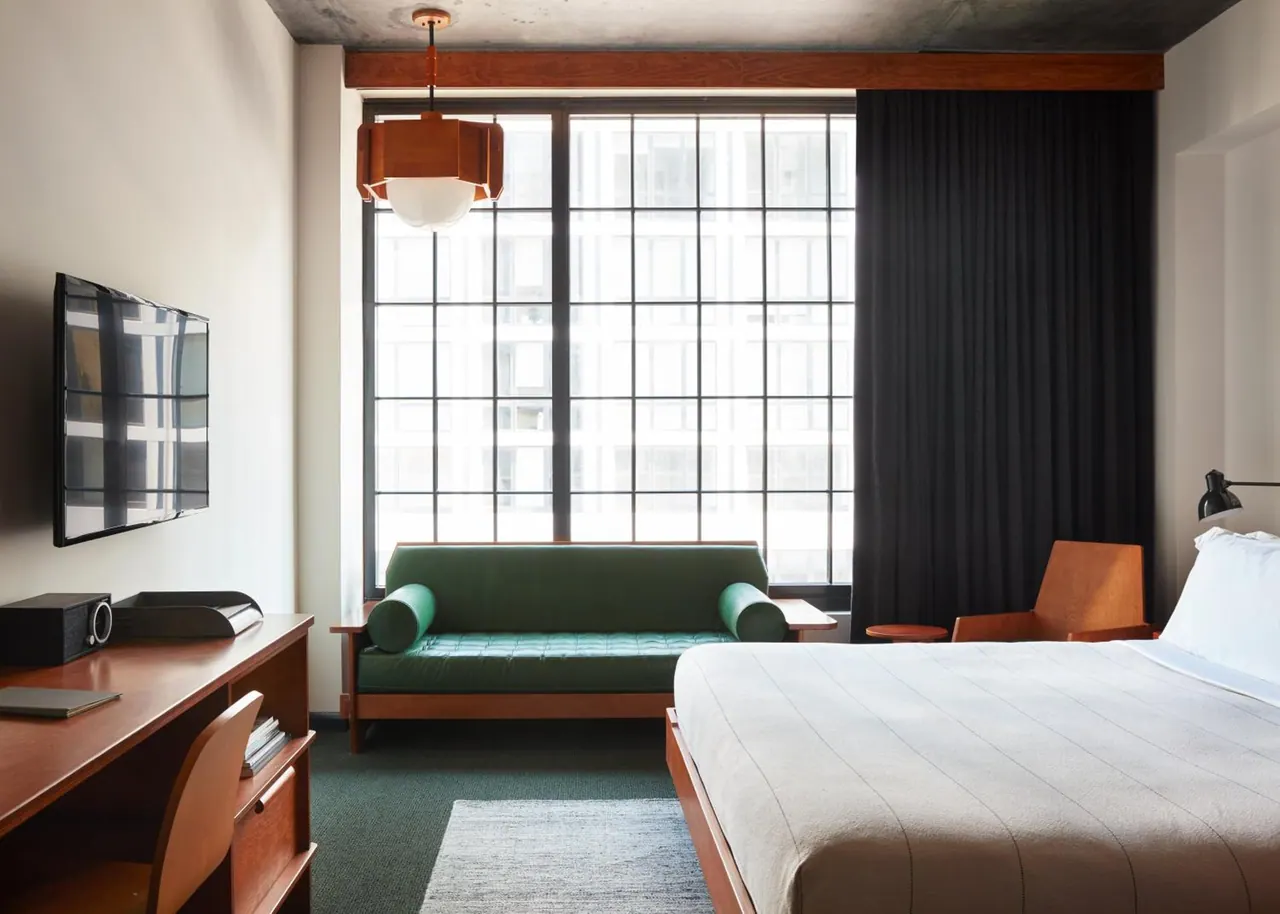 A hotel bedroom with a king bed, a green sofa beside a large grid-paned window, a wall-mounted TV, and a wooden desk with a chair.