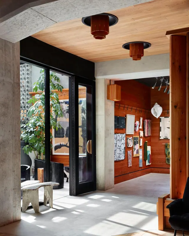 A lobby with concrete columns, a wood ceiling with pendant lights, a plant and concrete stool beside black glass doors, and a gallery wall with artworks.