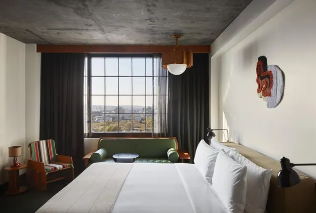 A king bed with white linen sits in a hotel room beside a green sofa, a striped armchair and a large window overlooking city rooftops.