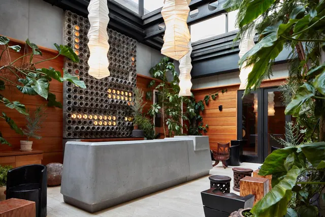 A lobby with a concrete reception desk, hanging paper lanterns, wood-panelled walls and plants beneath a skylit roof.