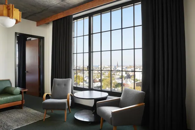 A seating area with two grey armchairs and a round black table sits beside a large window looking over city rooftops and a church steeple.