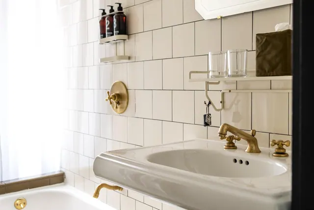 A white-tiled bathroom with a bathtub, a white sink with brass taps, and a wall shelf holding toiletry bottles and drinking glasses.