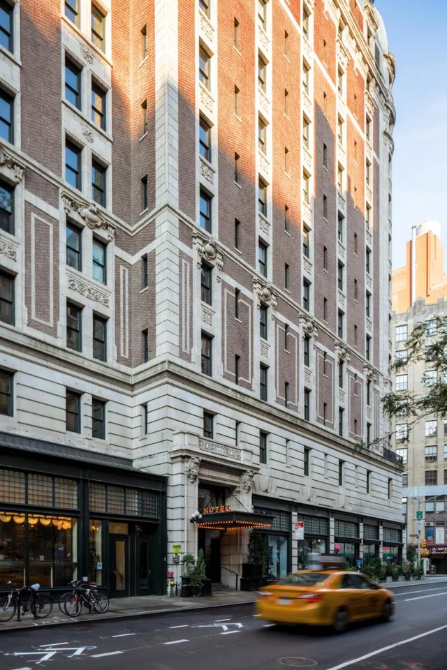 The exterior of a brick and stone hotel building with a lit HOTEL sign over the entrance, a blurred yellow taxi on the street, and bicycles parked on the sidewalk.