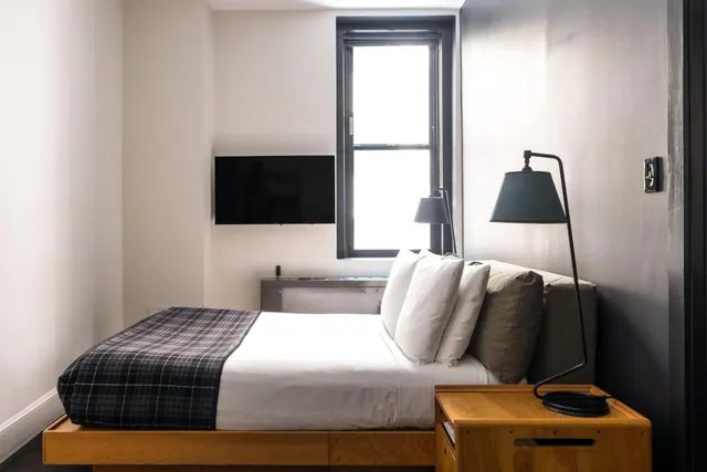 A bedroom with a platform bed under a plaid throw, a wall-mounted television, a window and a wooden nightstand with a lamp.
