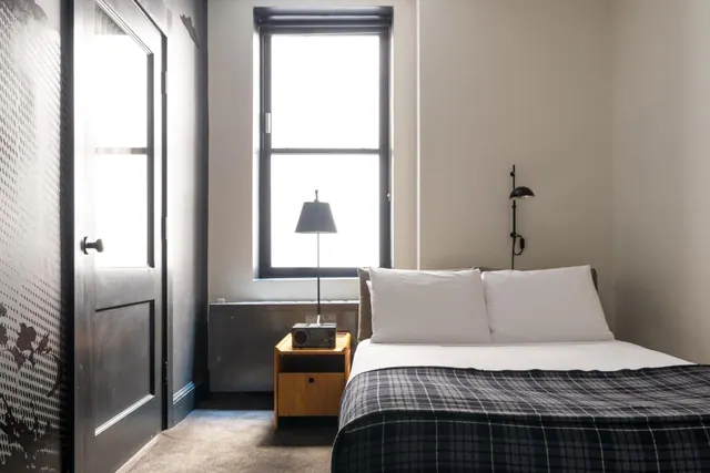 A bedroom with a double bed under a plaid blanket, a bedside table with a lamp and radio, a wall-mounted lamp, and a tall window letting in bright light.