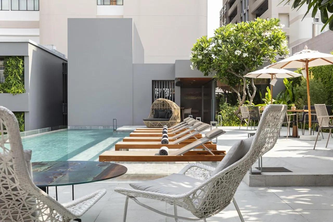 Wooden sun loungers, wicker chairs and cream parasols sit beside a rectangular pool with a frangipani tree and a grey building behind.
