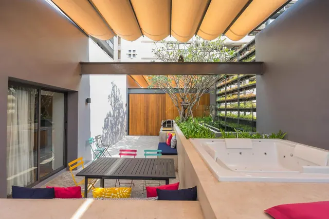 An outdoor terrace with a built-in hot tub, a dark metal table, colourful chairs and a fabric awning overhead, with a tree and planted wall beyond.