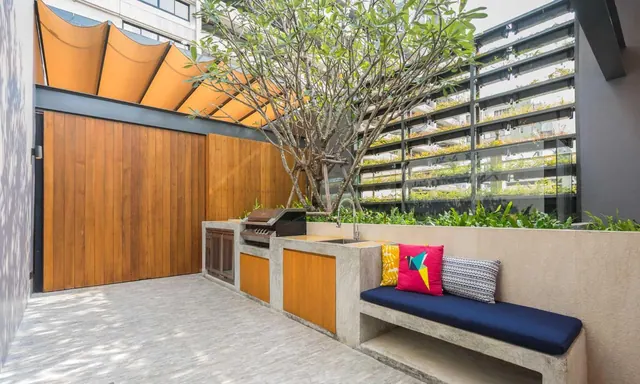 An outdoor terrace with a built-in barbecue grill and concrete counter, a concrete bench with a navy cushion and colourful pillows, a tree, and a building facade with planted balconies behind.