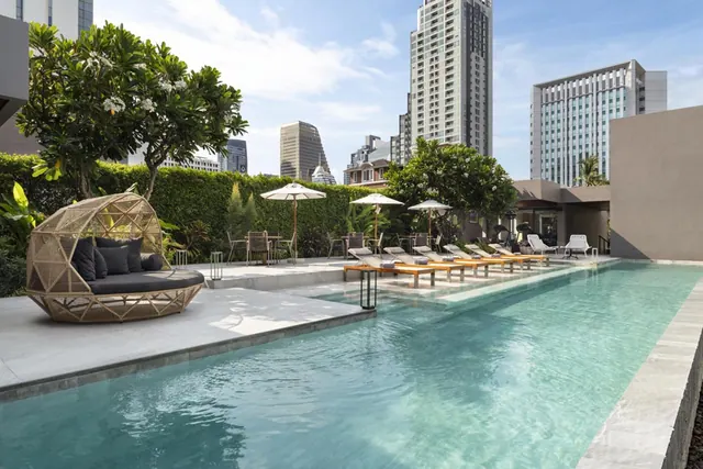 A long outdoor swimming pool with sun loungers, shade umbrellas, a wicker daybed and high-rise towers in the background.