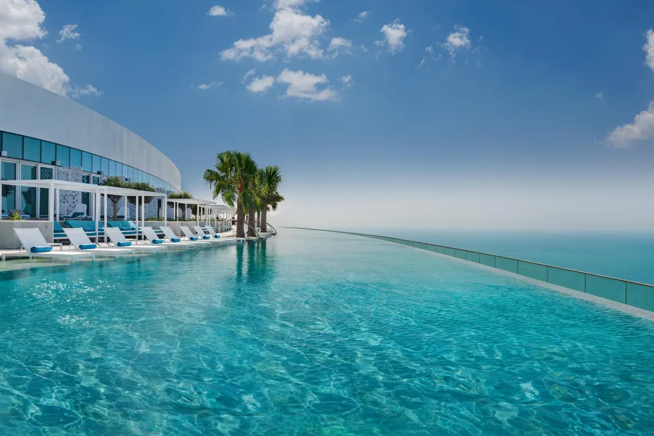 An infinity-edge pool with white sun loungers and palm trees overlooks the sea under a blue sky.