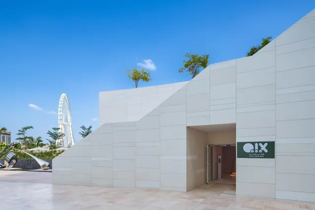A white stepped-facade building entrance with a green sign reading PLAY YOUR WAY, with a large Ferris wheel and a metal sculpture visible to the left under a clear blue sky.