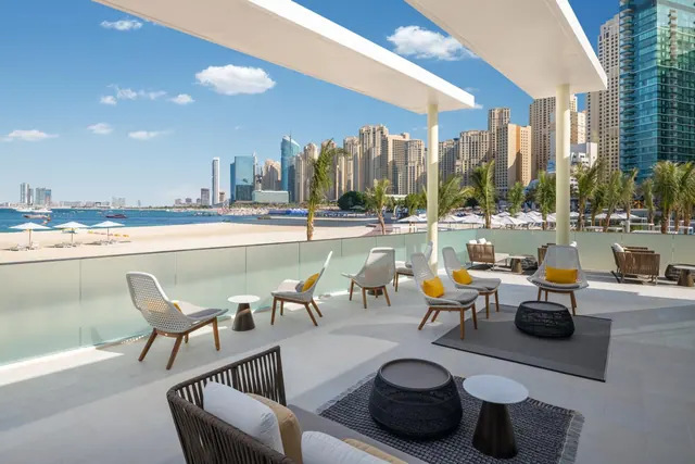 A covered terrace with lounge chairs and armchairs overlooks a sandy beach, the sea and a skyline of high-rise towers.