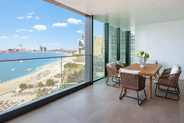 A furnished balcony with a wooden dining table and wicker chairs overlooks a sandy beach, the sea and a city skyline.