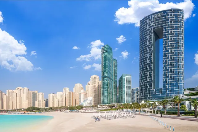 A sandy beach with white sun loungers and closed white umbrellas faces turquoise water and a row of high-rise towers, including a tall curved glass tower on the right.