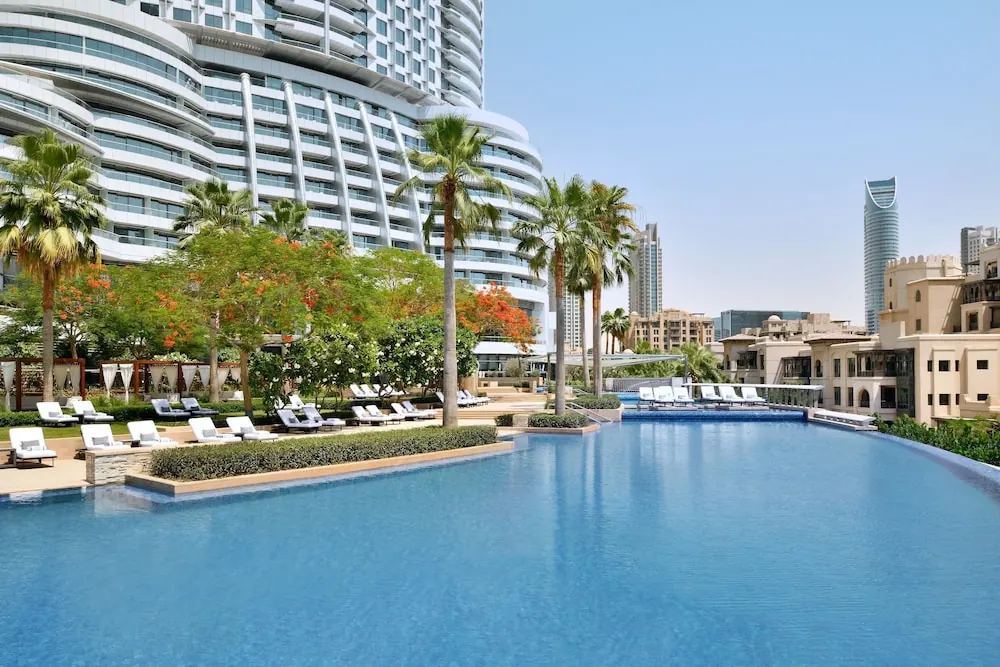 A large outdoor pool with white sun loungers along its edge, palm trees, a high-rise building and city towers visible behind.