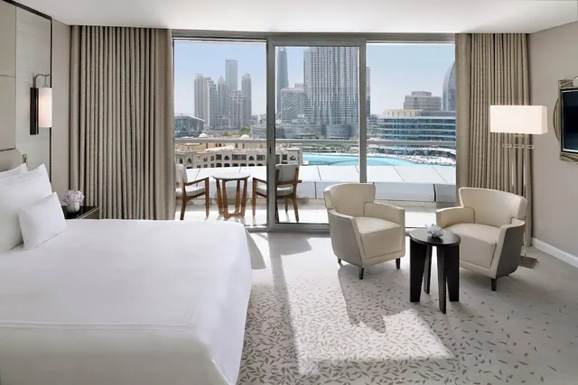 A bedroom with a made king bed, two armchairs and a sliding glass door opening onto a balcony with a view of city towers and a pool.