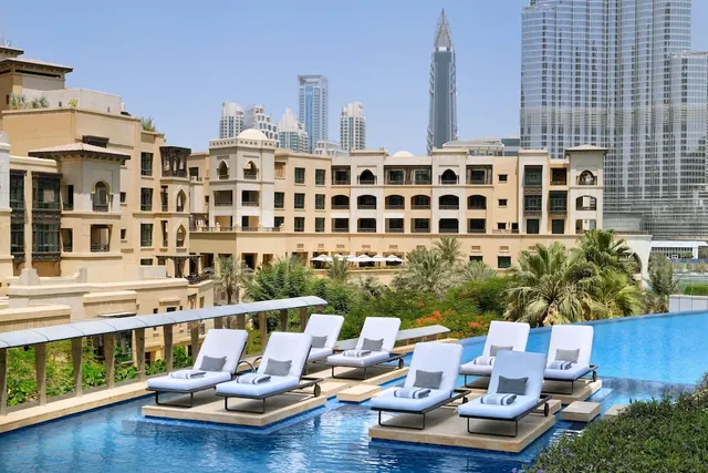 White-cushioned sun loungers sit in an infinity-edge pool, with palm trees, low-rise buildings and the Burj Khalifa tower in the background.