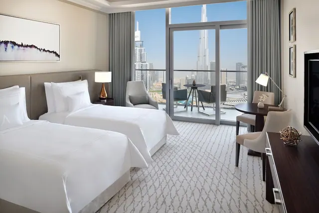 A bedroom with two made-up beds, an armchair, a desk and a wall-mounted television, with floor-to-ceiling windows and a balcony view of the Burj Khalifa.