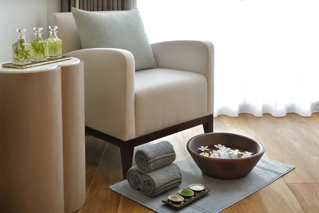 A cream armchair with a grey cushion sits beside a side table of glass bottles, with rolled towels and a wooden foot bowl on a grey mat in front.