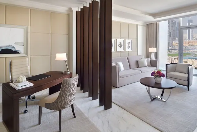 A suite living area with a wooden work desk, a grey sofa, an armchair and a round coffee table, with floor-to-ceiling windows showing city towers.