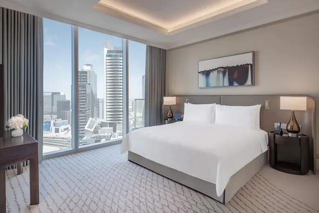 A king bed with white linen sits in a bedroom beside floor-to-ceiling windows that overlook a city skyline of high-rise towers.