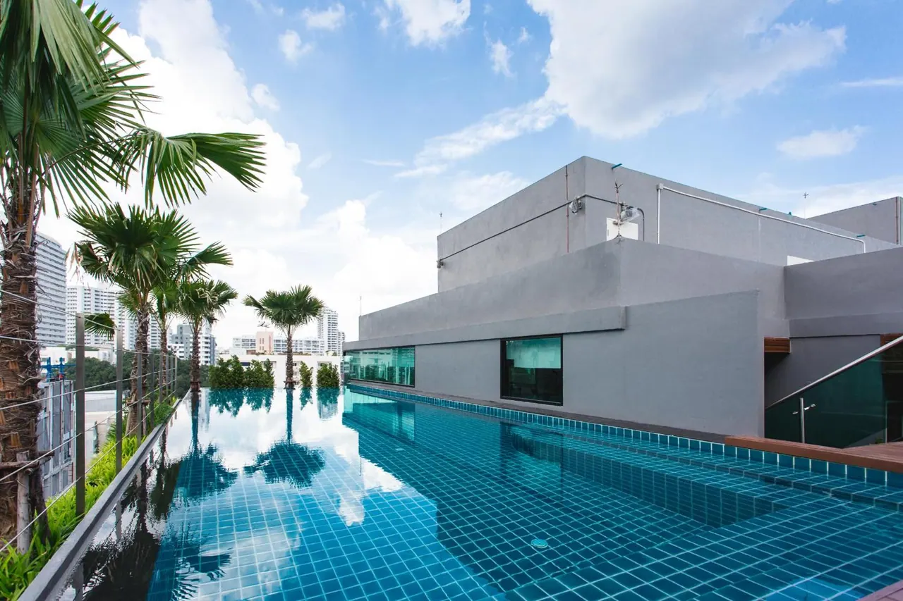 A rooftop swimming pool with blue tiles runs beside a grey building, with palm trees on the left and city towers in the background.