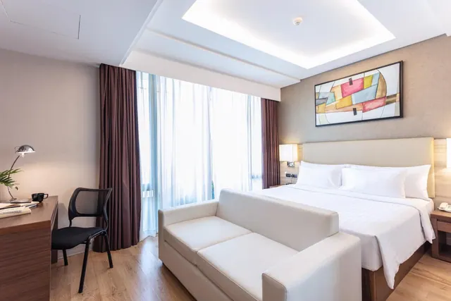 A hotel bedroom with a king bed, a cream sofa at the foot of the bed, a wooden desk and chair by the window, and an abstract painting above the headboard.