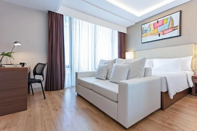 A hotel bedroom with a double bed, a white sofa, a desk with a lamp and chair, and large windows with sheer curtains.