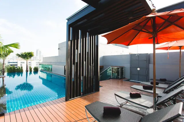 A rooftop swimming pool with a wooden deck, sun loungers with maroon towels, orange umbrellas, palm trees and city buildings in the background.