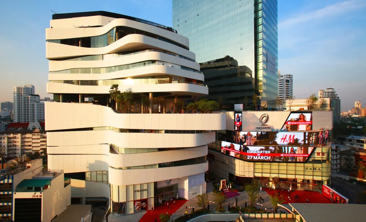 Exterior view of the EmQuartier mall with curved white terraces, large LED advertising screens and a red-carpeted plaza at street level.