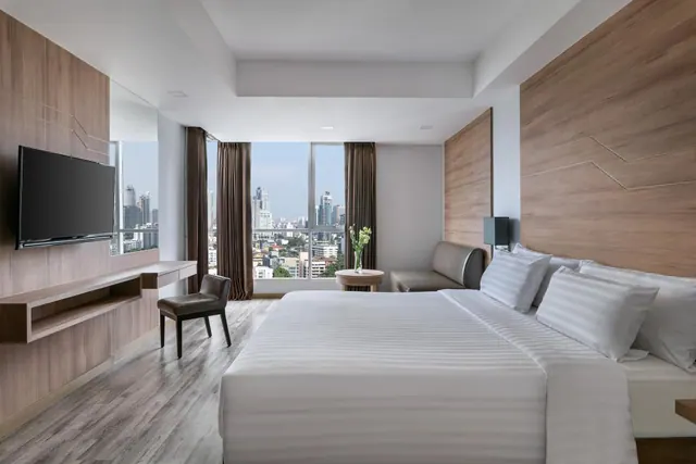 A hotel bedroom with a king bed, wall-mounted television, work desk and grey sofa, with floor-to-ceiling windows overlooking city high-rises.