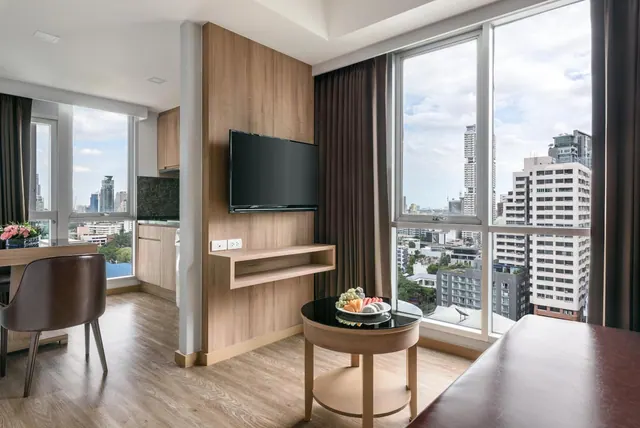 A living area with a wall-mounted television, a round side table with a fruit plate, a kitchenette and floor-to-ceiling windows overlooking high-rise buildings.