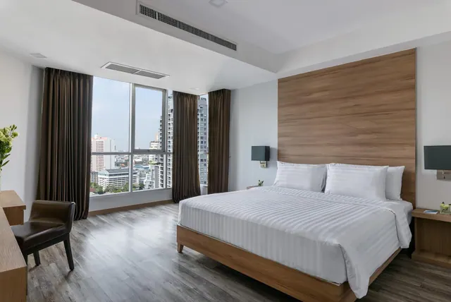A bedroom with a large made bed, a leather chair, and floor-to-ceiling windows looking out over high-rise buildings.