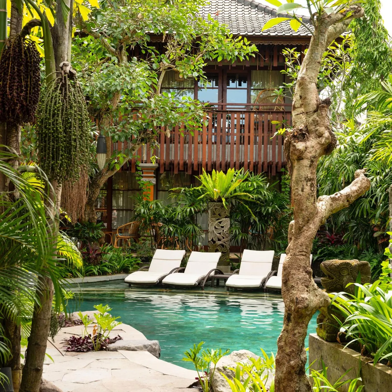 A turquoise pool with white sun loungers sits among tropical plants beside a traditional Balinese wooden building with a tiled roof.