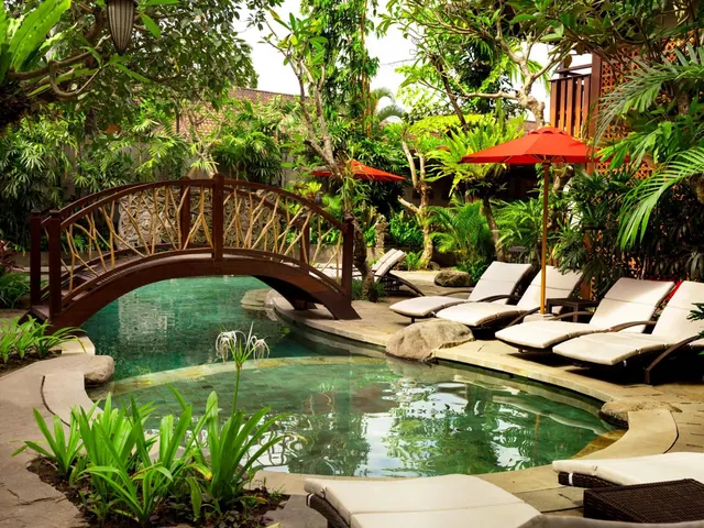 A kidney-shaped pool with a wooden arched footbridge, white sun loungers and red umbrellas sits among tropical garden plants.