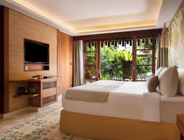 A bedroom with a made king bed, a wall-mounted television and a wooden cabinet, with sliding doors opening onto lush tropical greenery.