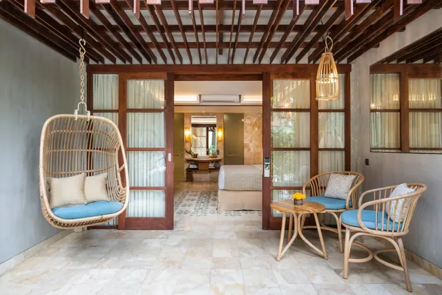 A covered terrace with a hanging rattan chair, two rattan armchairs and a small round table, with glass doors open to a bedroom with a bed and a wall air conditioner.