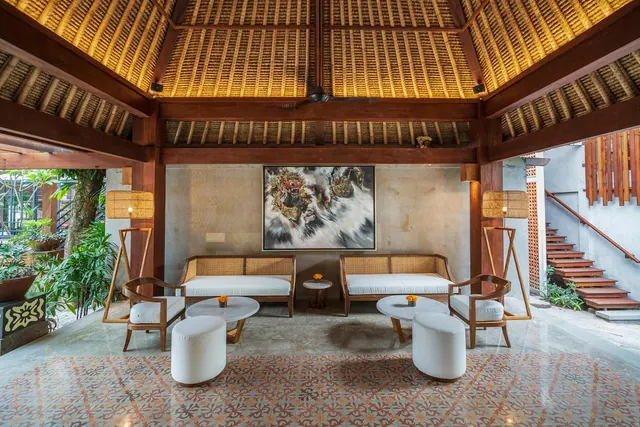 A lounge seating area in a hotel lobby with rattan sofas, round marble tables, white ottomans and a large painting under a thatched timber roof.
