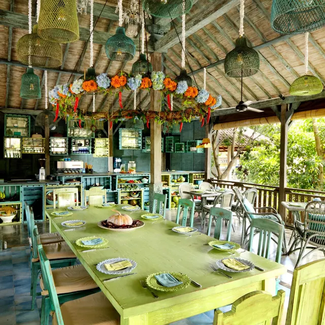 A long green dining table set with plates and napkins sits under woven pendant lamps and a floral garland in an open-sided restaurant, with trees visible beyond the railing.