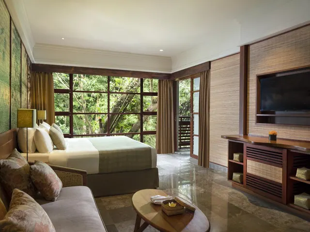 A bedroom with a king bed, a sofa, a wall-mounted television and a wooden cabinet, with a large window and glass door looking onto dense green trees.