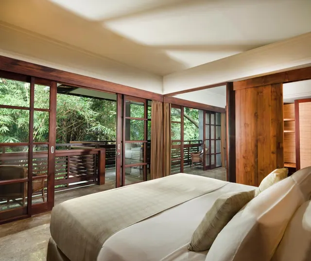 A bedroom with a made bed faces wooden-framed glass doors that open onto a balcony with wicker chairs and a view of green foliage.