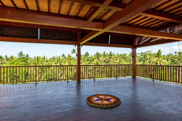 A covered wooden terrace with a floral offering on the deck looks out over palm trees and greenery under a clear sky.
