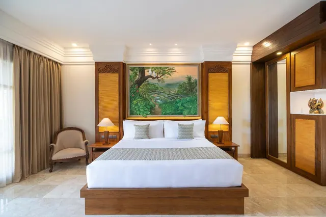 A bedroom with a king bed beneath a large painting of a green landscape, with an armchair by the curtained window and a wooden wardrobe on the right.