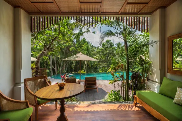 A covered terrace with a round wooden table, cane chairs and a green daybed looks out onto a swimming pool and tropical greenery.